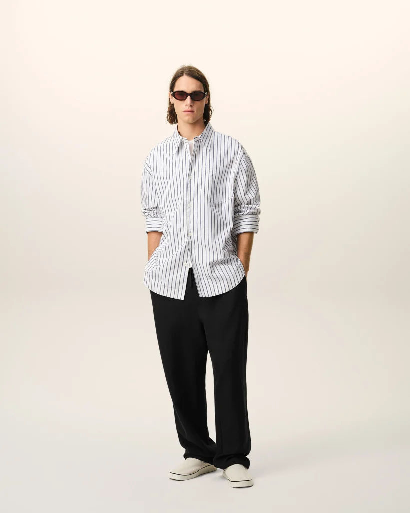 Person standing against a light background, wearing a loose striped button-down shirt, black pants, white sneakers, and dark sunglasses.