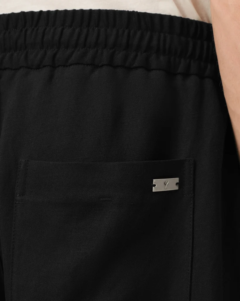 Close-up of black pants showing an elastic waistband, textured fabric, and a small metal logo tag on the pocket.