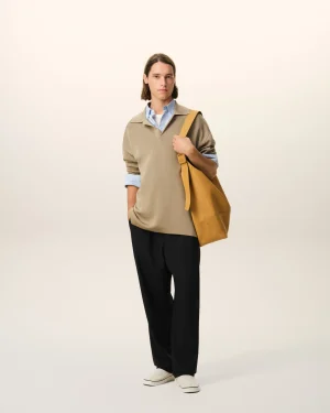 Man in a beige sweater over a blue shirt, black trousers, white sneakers, carrying a tan shoulder bag against a pale gradient background.