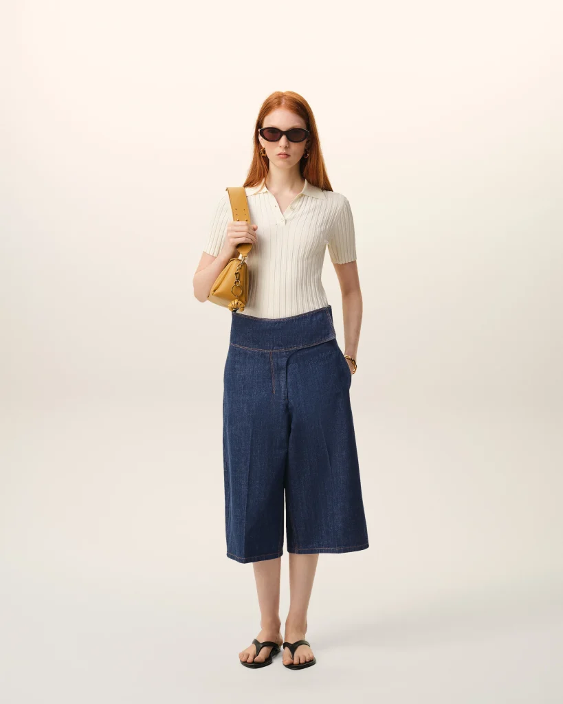 Woman standing and facing forward in a white ribbed polo, dark wide-leg denim skirt, black sandals, sunglasses, and a yellow shoulder bag.