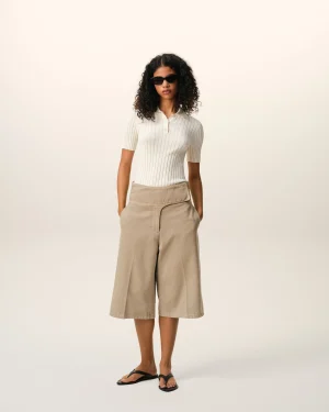 Full-body shot of a woman in a white ribbed polo shirt, beige wide-leg shorts, black sandals, and sunglasses against a light backdrop.