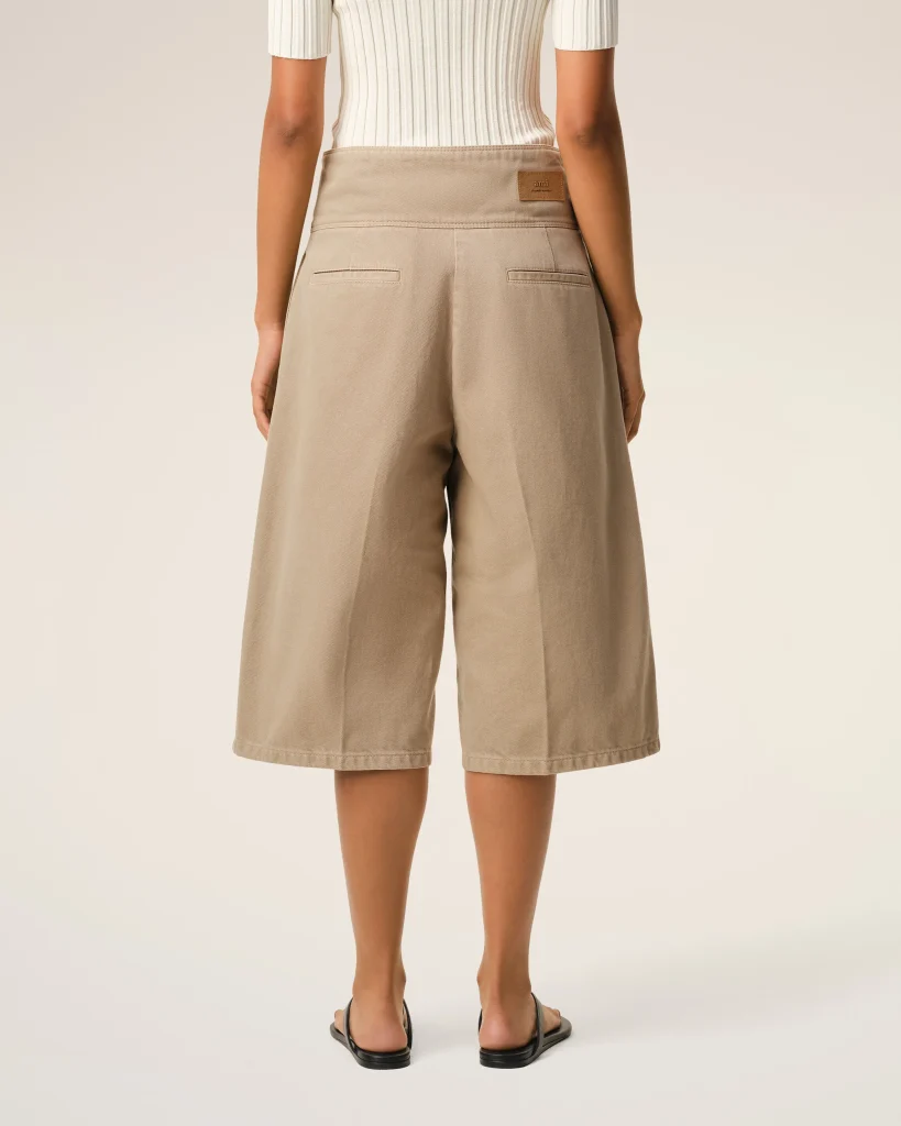 Back view of a person wearing beige wide-leg shorts, a white ribbed top, and black sandals against a neutral background.