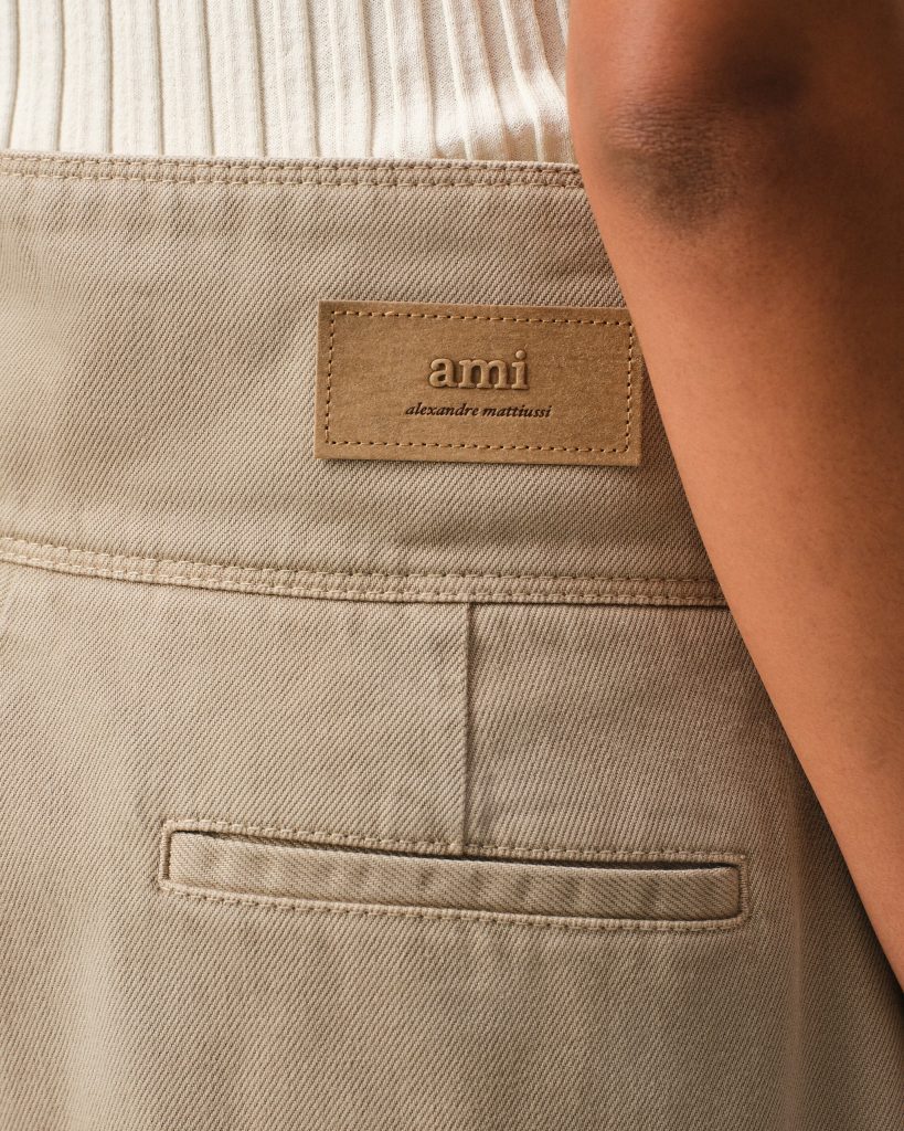 Beige denim jacket pocket with a tan leather brand patch reading 'ami alexandre mattiussi' on it.