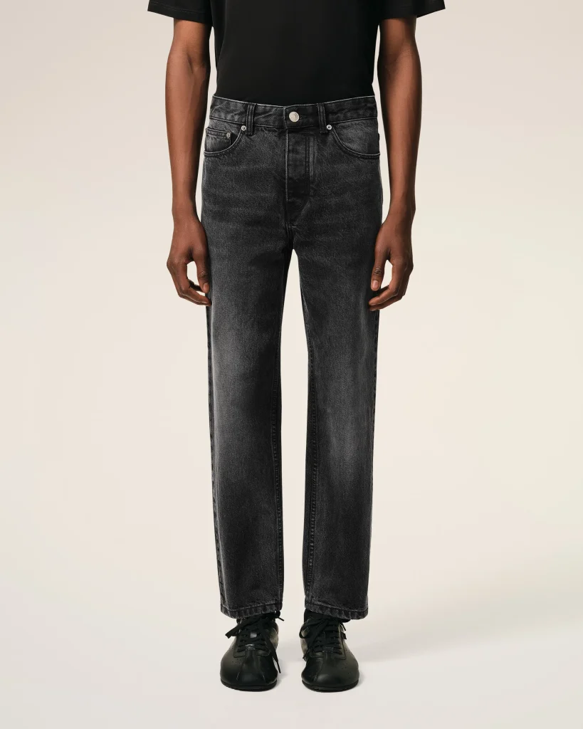 Person wearing a black T-shirt with dark gray jeans and black sneakers, shown from waist to feet against a beige background.