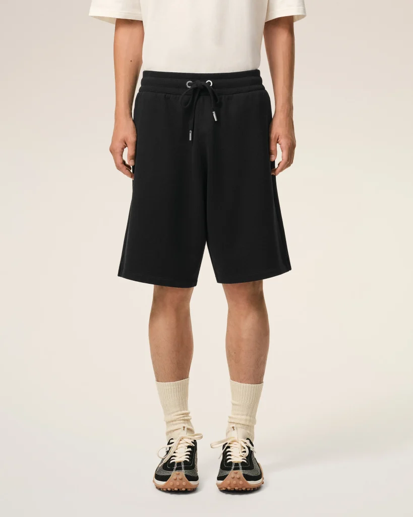 Lower half of a person wearing black drawstring shorts, a white T-shirt, cream socks, and black sneakers, standing against a light background.