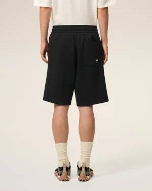 Back view of a person wearing black shorts with a rear pocket, white shirt, beige socks, and black sandals with tan soles.