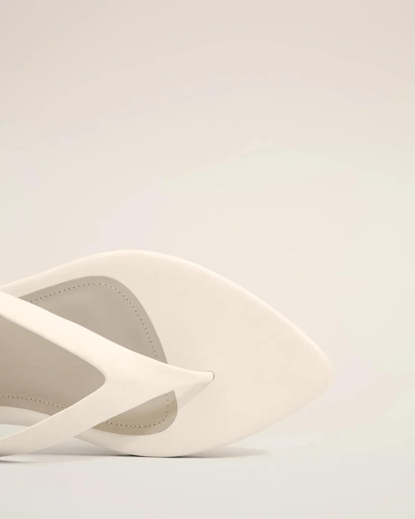 White sandal close-up showing leather straps and stitching on a pale background.