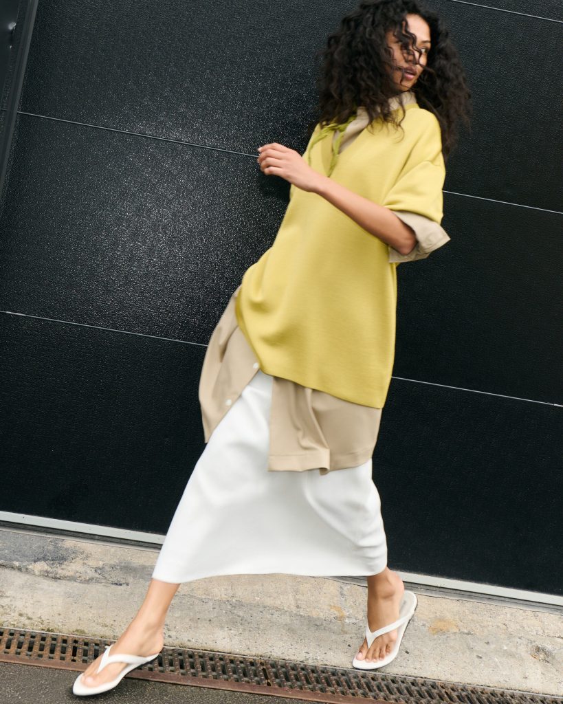 Person walking past a dark textured wall, wearing a pale yellow coat over beige clothing and white sandals, captured mid-step on a sidewalk