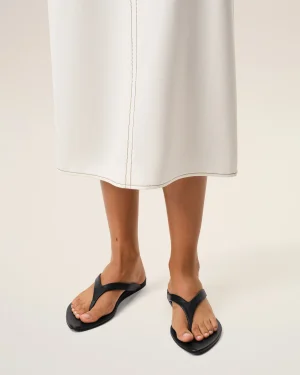 Lower half of a person wearing a white knee-length skirt with brown stitching and black sandals at the feet.