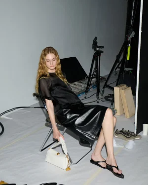 A woman in a black satin dress sits in a photography studio among lighting stands and equipment, with a white handbag on the floor nearby.