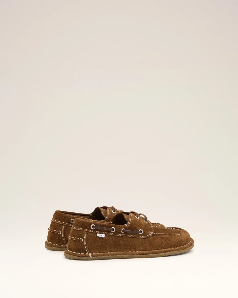 Brown suede boat shoes with white stitching and laces on a pale background.