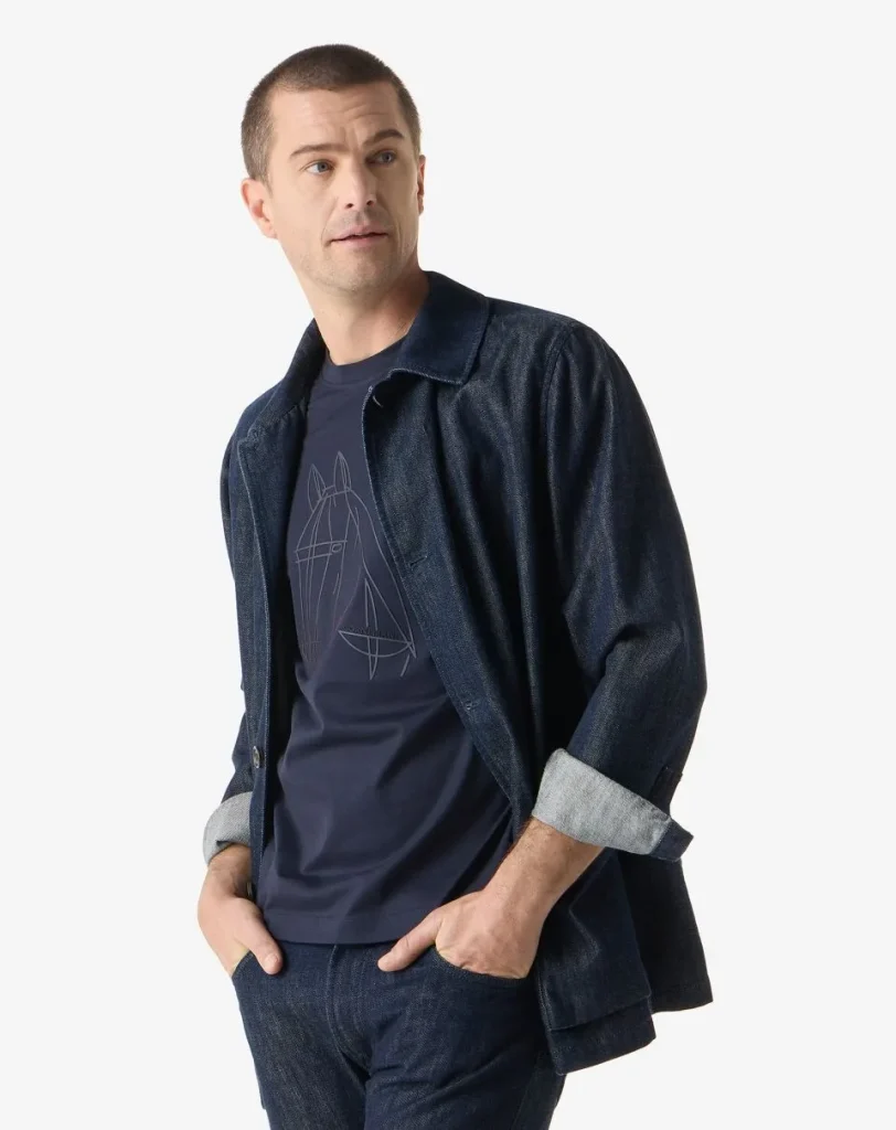 Man wearing a dark denim jacket and jeans with a navy T-shirt featuring a geometric horse graphic, hands in pockets, looking to the side.