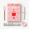 Pink cupcake decorating book with a variety of icing tools and accessories laid out: spatula, piping tips, parchment roll tied with pink ribbon, and pink cupcake liners.