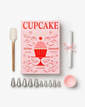 Pink cupcake decorating book with a variety of icing tools and accessories laid out: spatula, piping tips, parchment roll tied with pink ribbon, and pink cupcake liners.