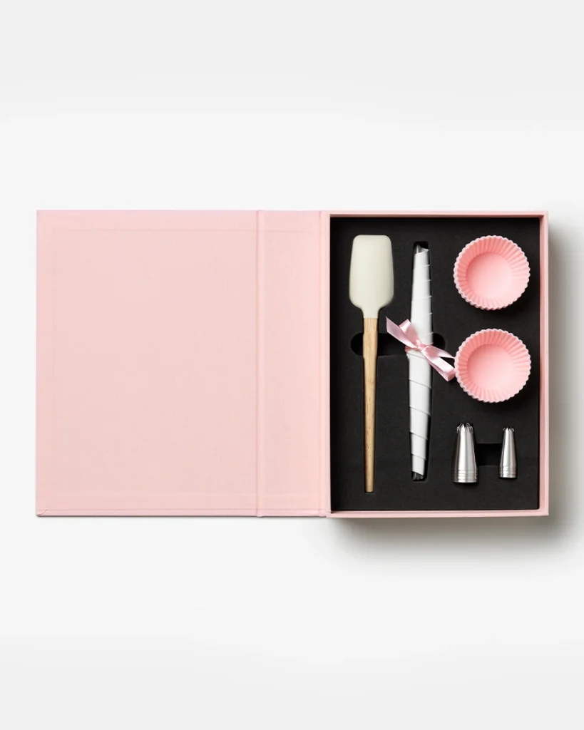 Pink gift box opened to reveal a cake decorating kit: spatula, piping bag with ribbon, two pink cupcake liners, and metal decorating tips in a black insert