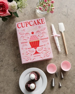 Pink cupcake cookbook titled 'Cupcake Tips' on a gray surface with decorating tools and small cupcakes on a white plate.