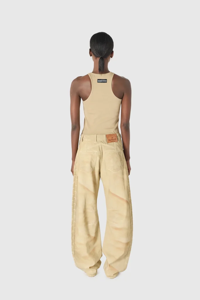 Back view of a person wearing a beige sleeveless top and wide beige pants with a low ponytail and a small back patch reading 'Marine Serre'.