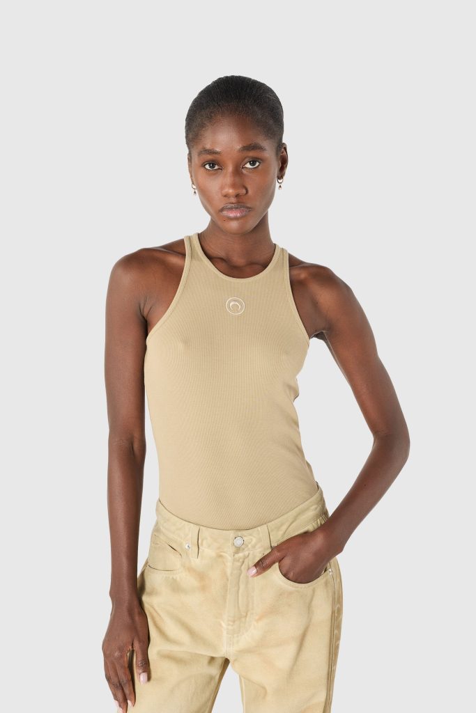Model wearing a beige sleeveless top and light tan cargo-style pants, standing against a light gray background with a neutral expression.