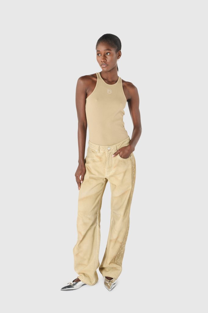 Woman in a beige sleeveless top and light tan pants stands against a gray background, looking to the left.