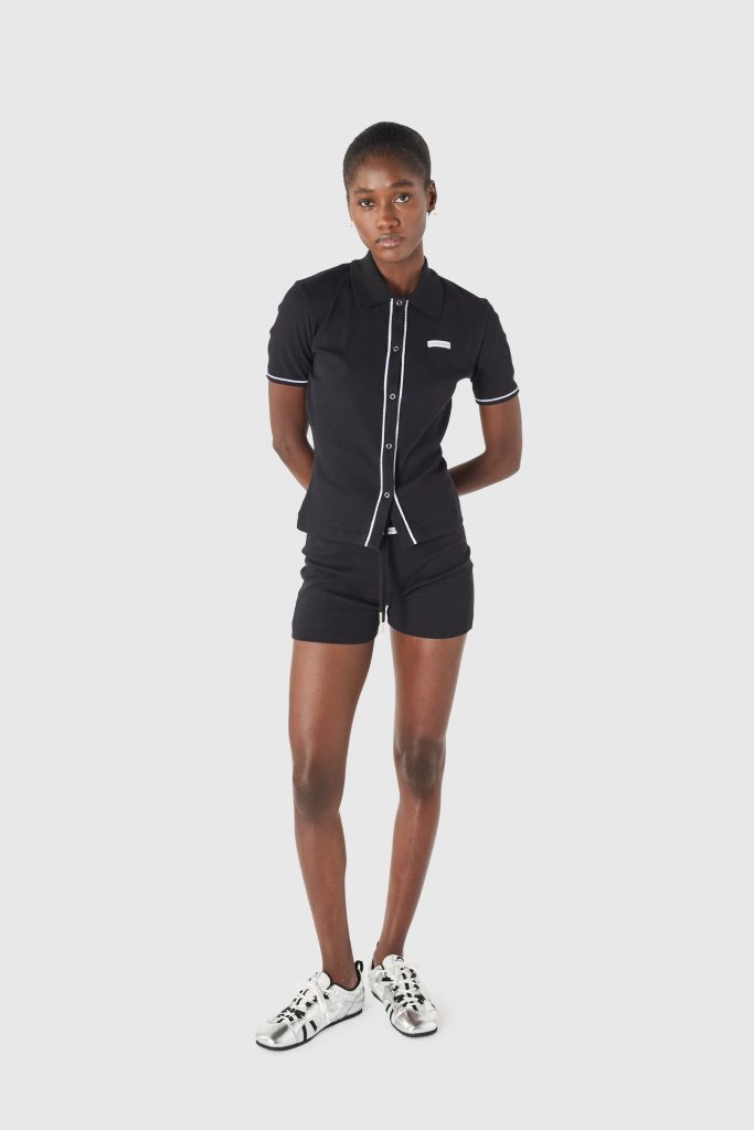 Full-body portrait of a woman in a black polo shirt with white piping and matching shorts, standing with hands behind her back against a light gray background.