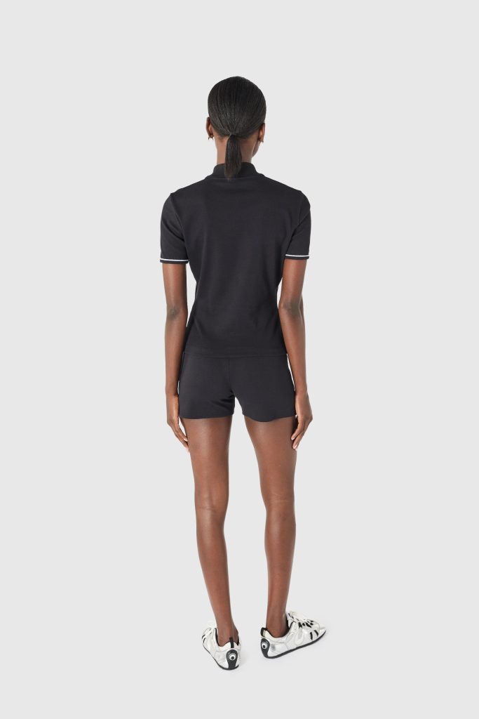 Back view of a person wearing a black polo shirt and black shorts, white sneakers, standing against a light gray background with arms at sides.