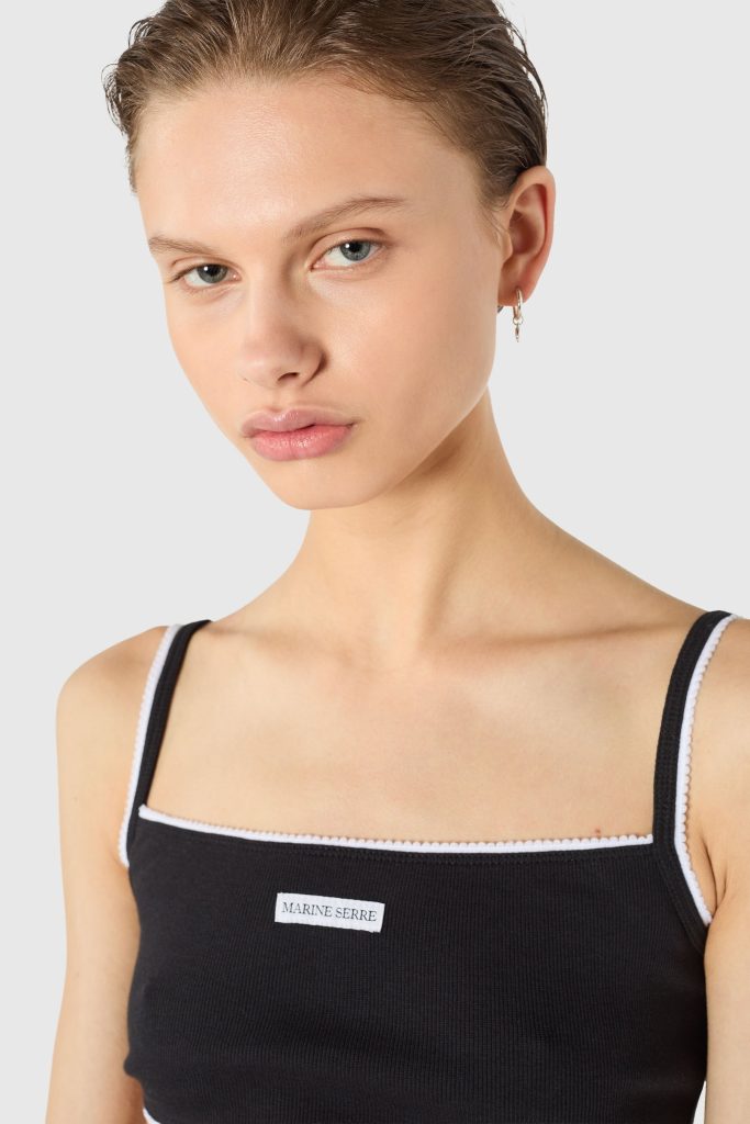 Close-up portrait of a woman wearing a black ribbed tank top with white trim and a small Marine Serre label on the chest, wearing a hoop earring.