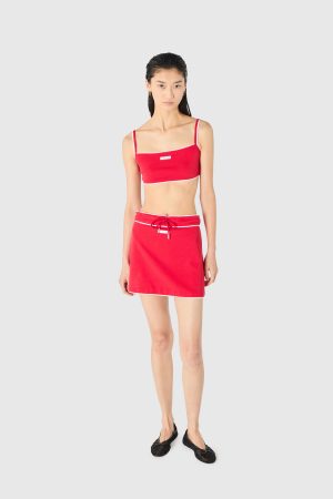 Young woman wearing a red bikini top with thin white-trim straps and a matching red skirt, standing against a light gray background.