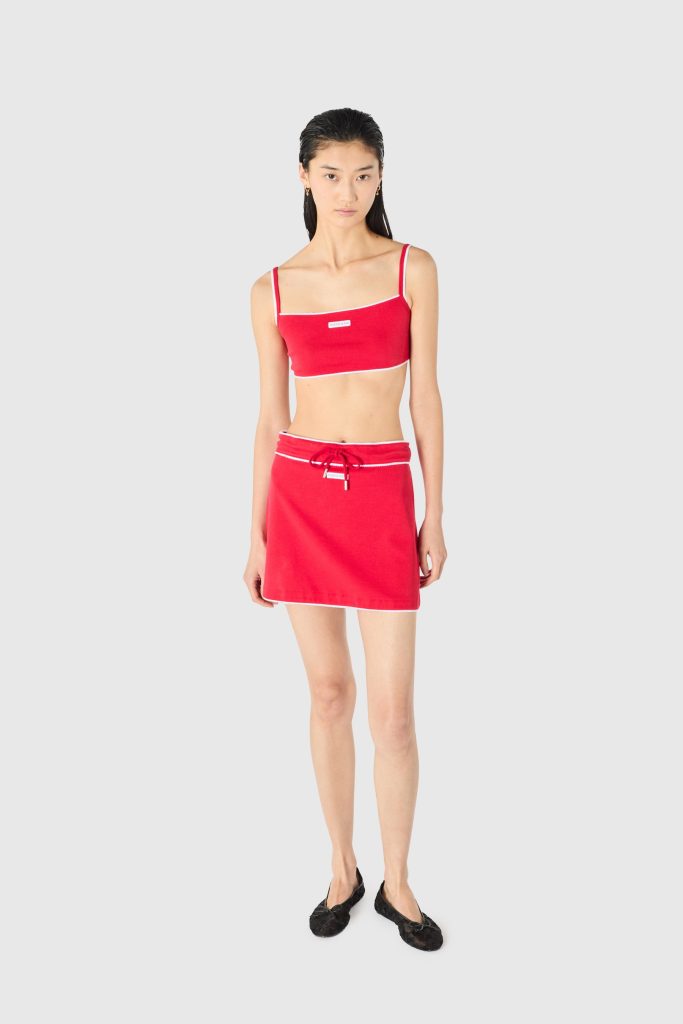 Young woman wearing a red bikini top with thin white-trim straps and a matching red skirt, standing against a light gray background.