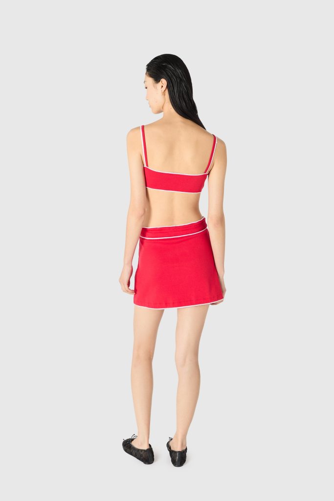 Rear view of a woman in a red bikini top and matching red skirt with white trim, standing against a light gray background, black shoes visible at the feet.