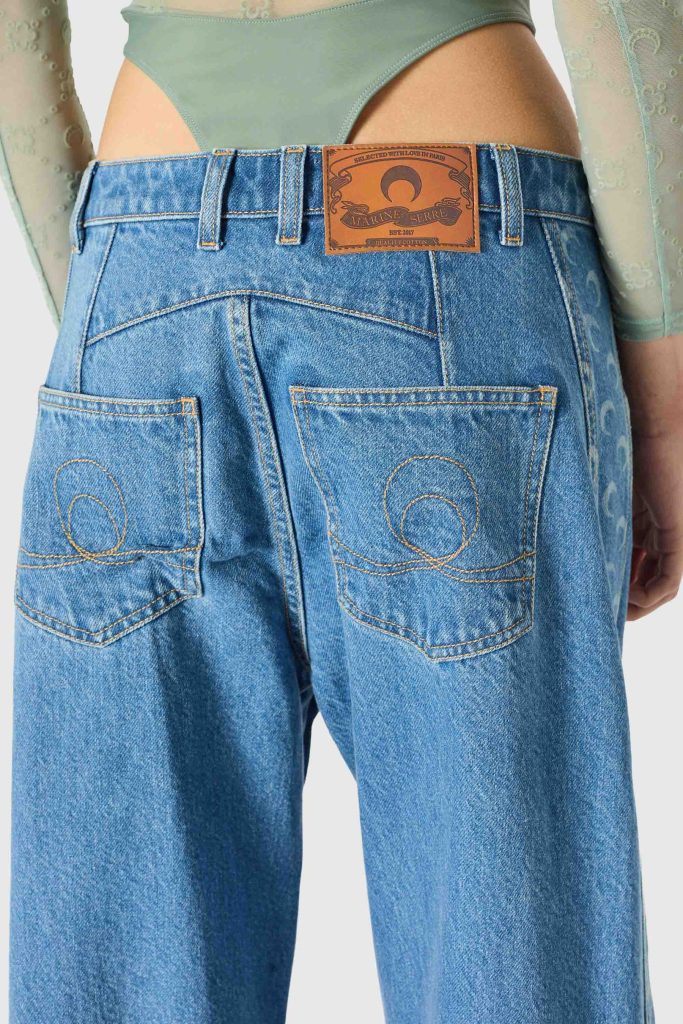 Rear view of a person wearing high-waisted blue jeans with two back pockets and a brown leather brand patch on the waistband; a green top peeks above the jeans.