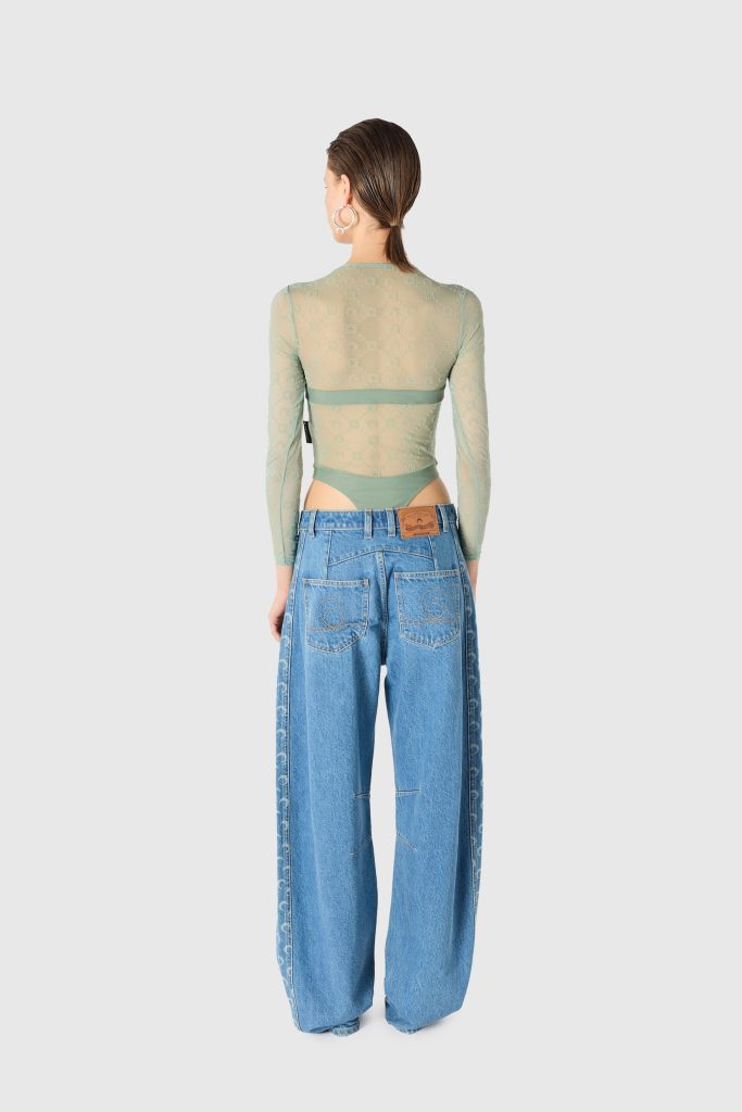 Back view of a person in a sheer mint green long-sleeve top and high-waisted blue jeans, hair tied at the nape, wearing large hoop earrings.
