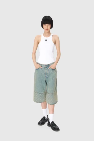 Woman standing in a white sleeveless top and patchwork denim skirt with hands in pockets against a light gray background.