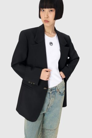 Woman in a black blazer over a white T-shirt and blue jeans, standing with hands on her hips against a gray background.