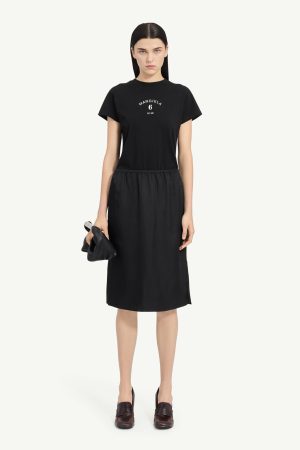 Model in an all-black outfit: short-sleeve black T-shirt tucked into a gathered black midi skirt, holding a small black clutch.