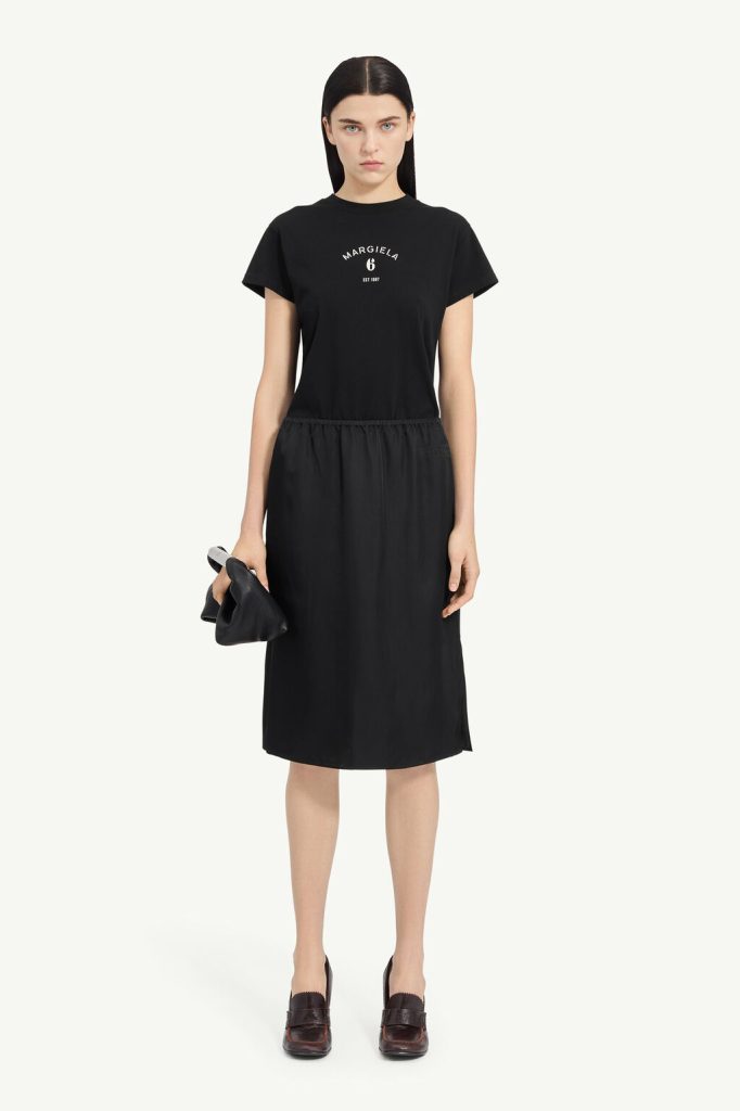 Model in an all-black outfit: short-sleeve black T-shirt tucked into a gathered black midi skirt, holding a small black clutch.