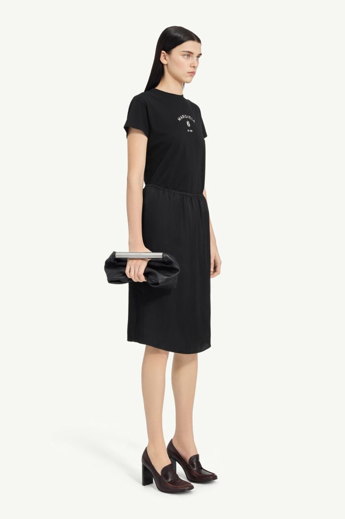 Model in a black short-sleeve midi dress, holding a black clutch, wearing burgundy heeled loafers against a light backdrop.