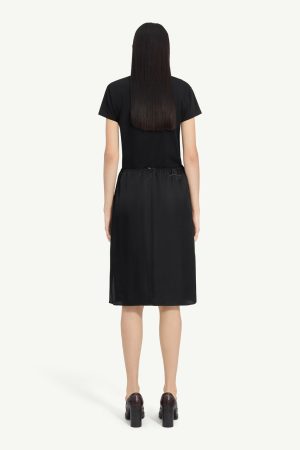Back view of a model wearing a black short-sleeve midi dress with an elastic waist and dark heeled shoes.
