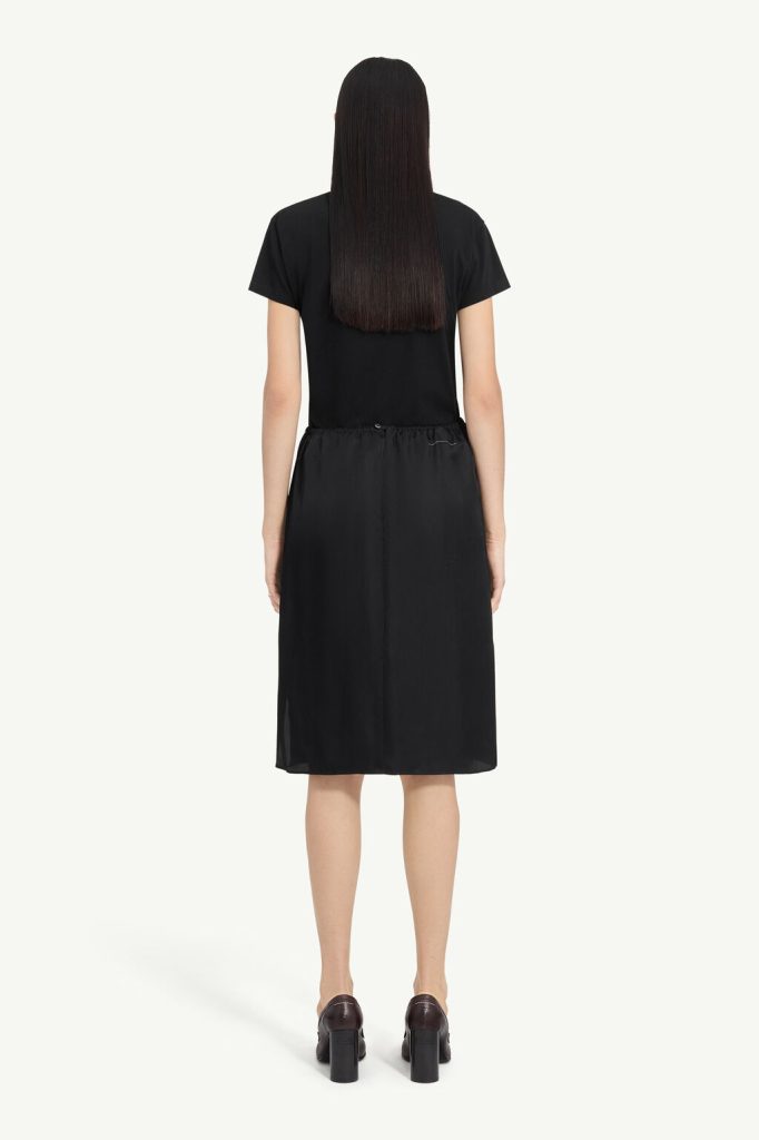 Back view of a model wearing a black short-sleeve midi dress with an elastic waist and dark heeled shoes.