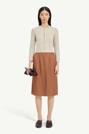Woman standing and facing camera in a beige textured cardigan and rust-brown midi skirt, holding a black clutch and wearing dark loafers against a pale background.
