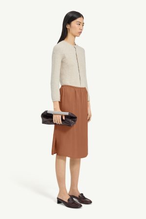 Side-profile of a woman wearing a cream textured cardigan, rust midi skirt, and a dark brown clutch with a silver bar, paired with dark loafers.