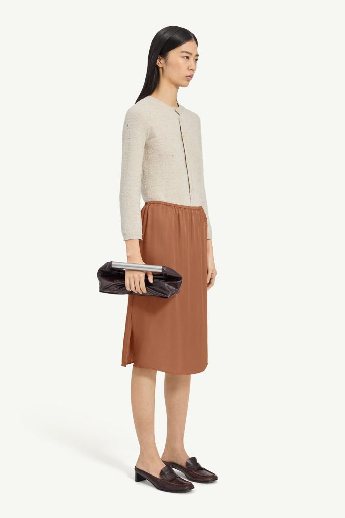 Side-profile of a woman wearing a cream textured cardigan, rust midi skirt, and a dark brown clutch with a silver bar, paired with dark loafers.