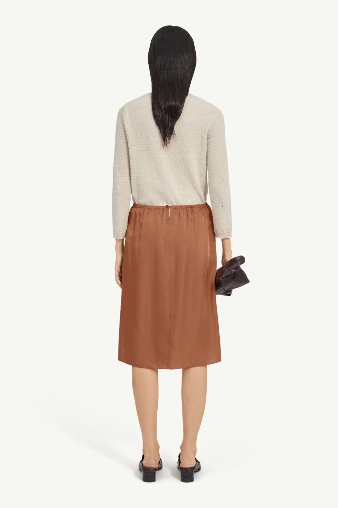 Back view of a person wearing a beige textured sweater and brown midi skirt, holding a dark bag at the side.