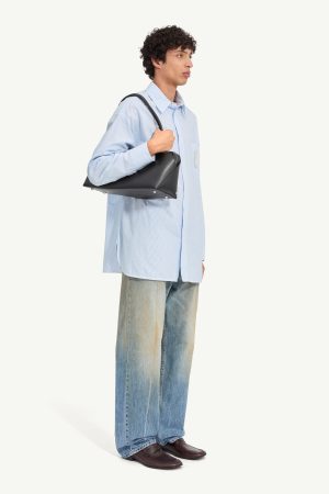 Young man in a light blue oversized shirt and faded jeans, carrying a black shoulder bag in profile orientation.