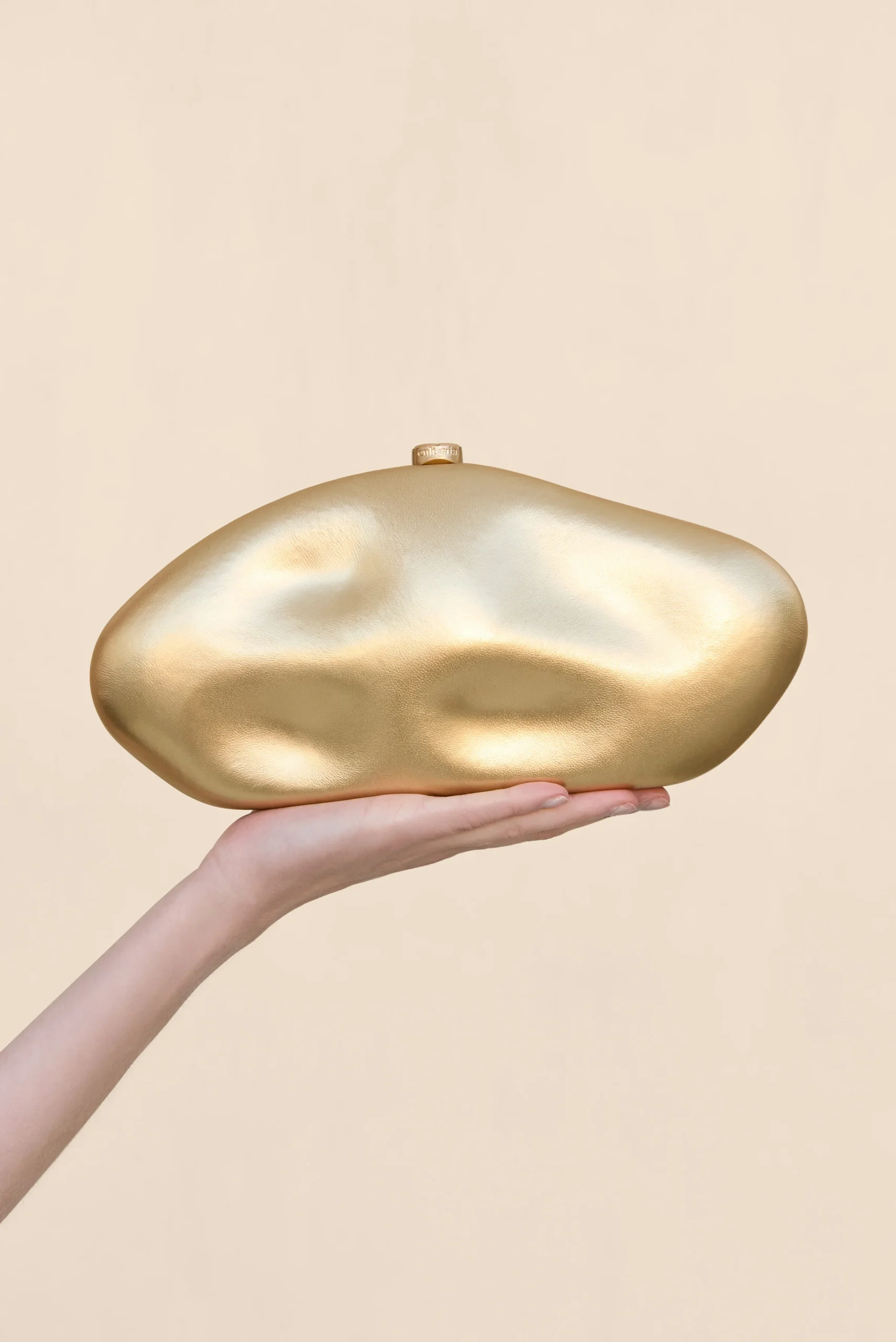 IMG_1264 - New Trend A hand holding a smooth, irregularly shaped gold sculpture against a pale beige background.