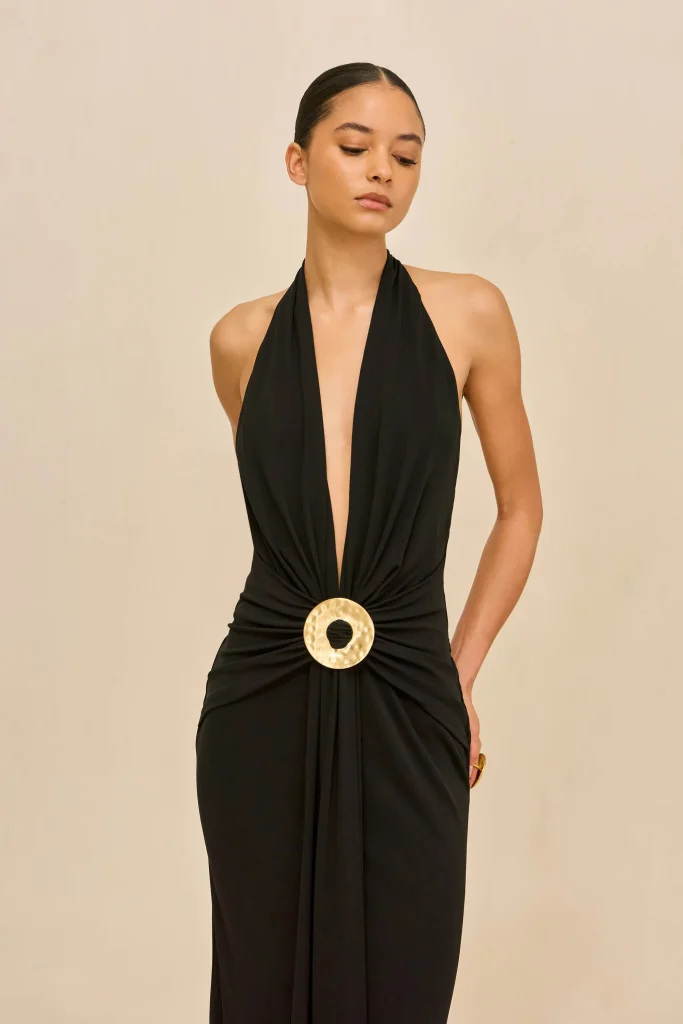 Woman in a black halter dress with a large circular gold belt, standing against a beige backdrop and looking downward.