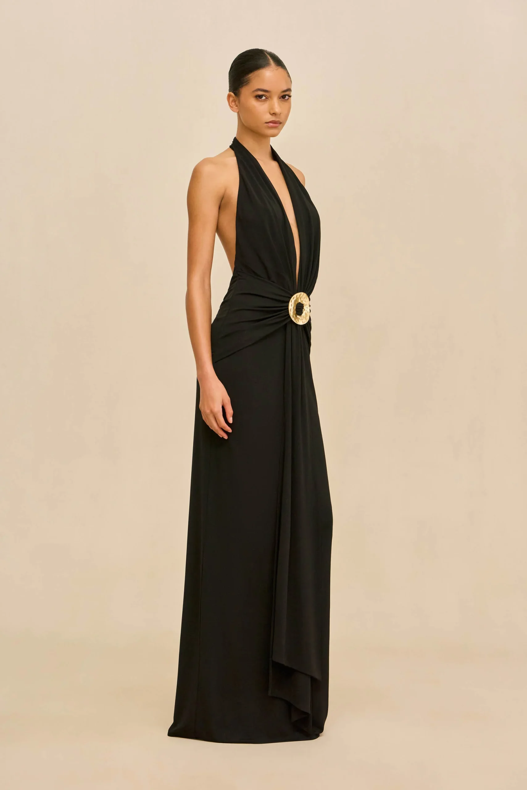 IMG_1279 - New Trend Full-length black halter gown with a deep V-neck and a gold circular belt buckle, standing against a beige backdrop.