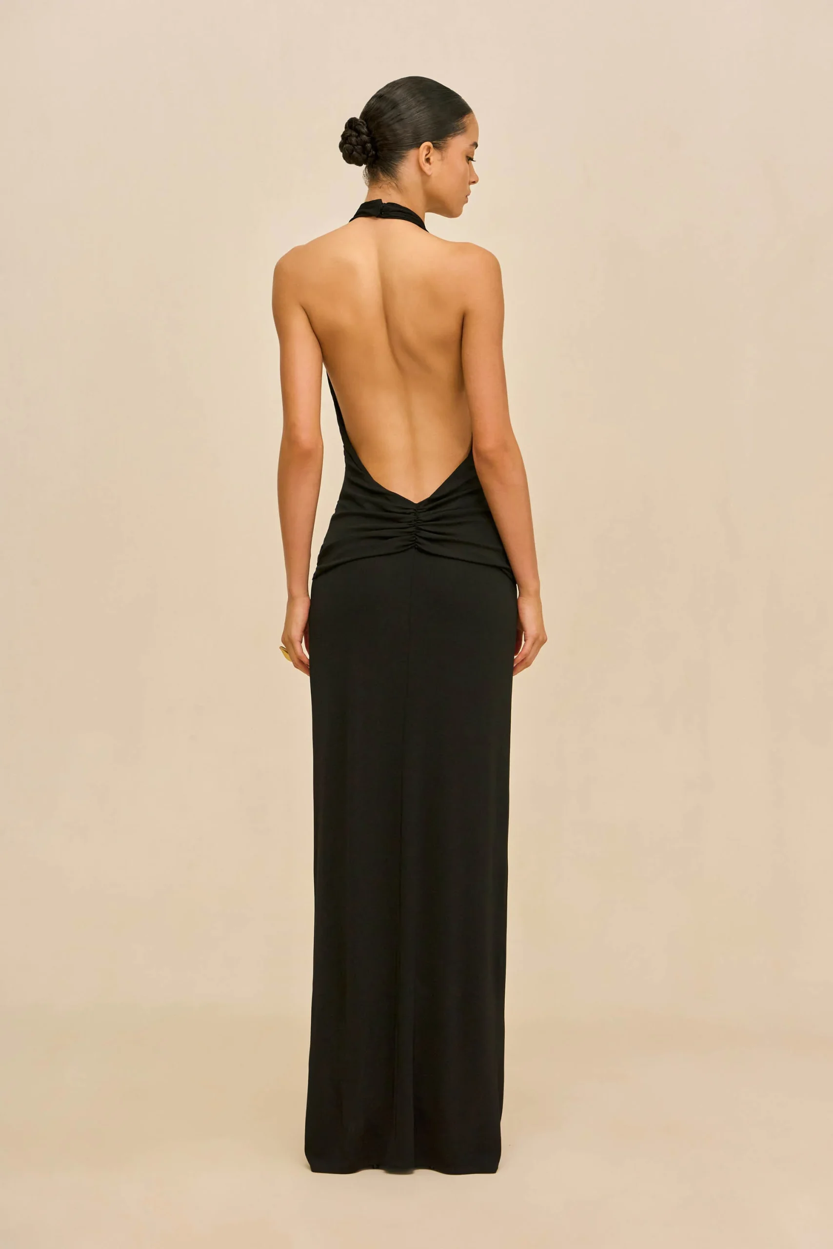 IMG_1280 - New Trend Back view of a woman in a black halter-back gown with a bun, standing against a beige backdrop.
