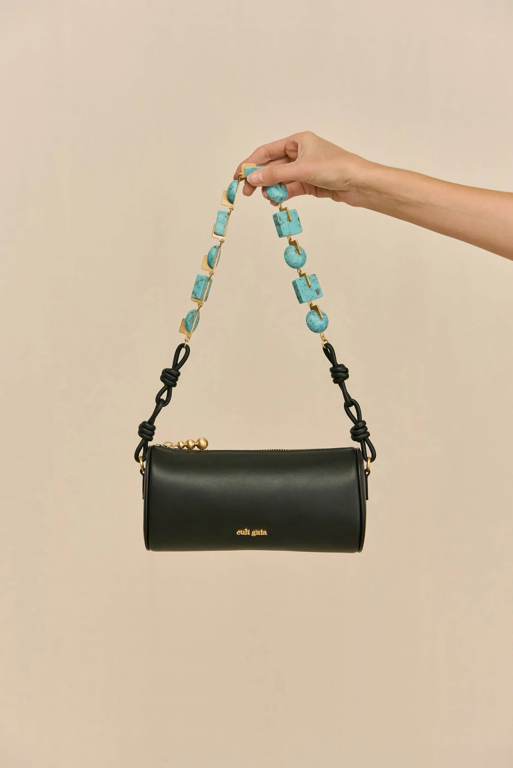 IMG_1290 - New Trend Hand holding a turquoise beaded strap above a black leather handbag against a beige background.