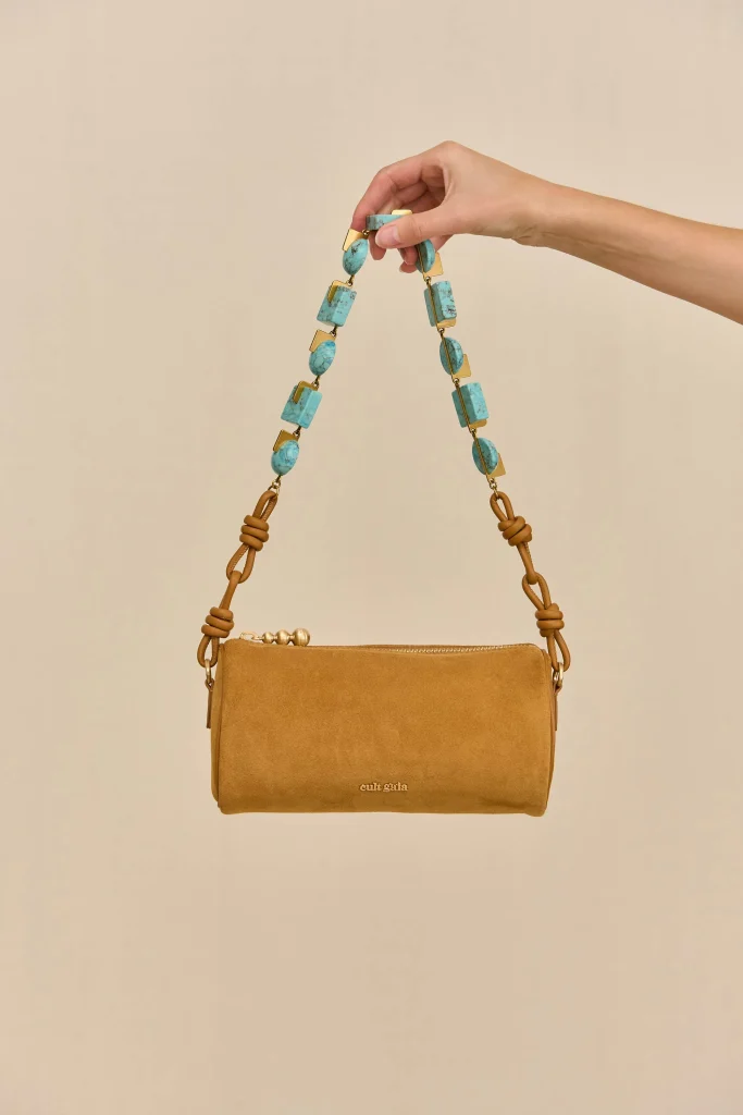 Beige Cult Gaia handbag with a turquoise beaded strap being held up by a hand.