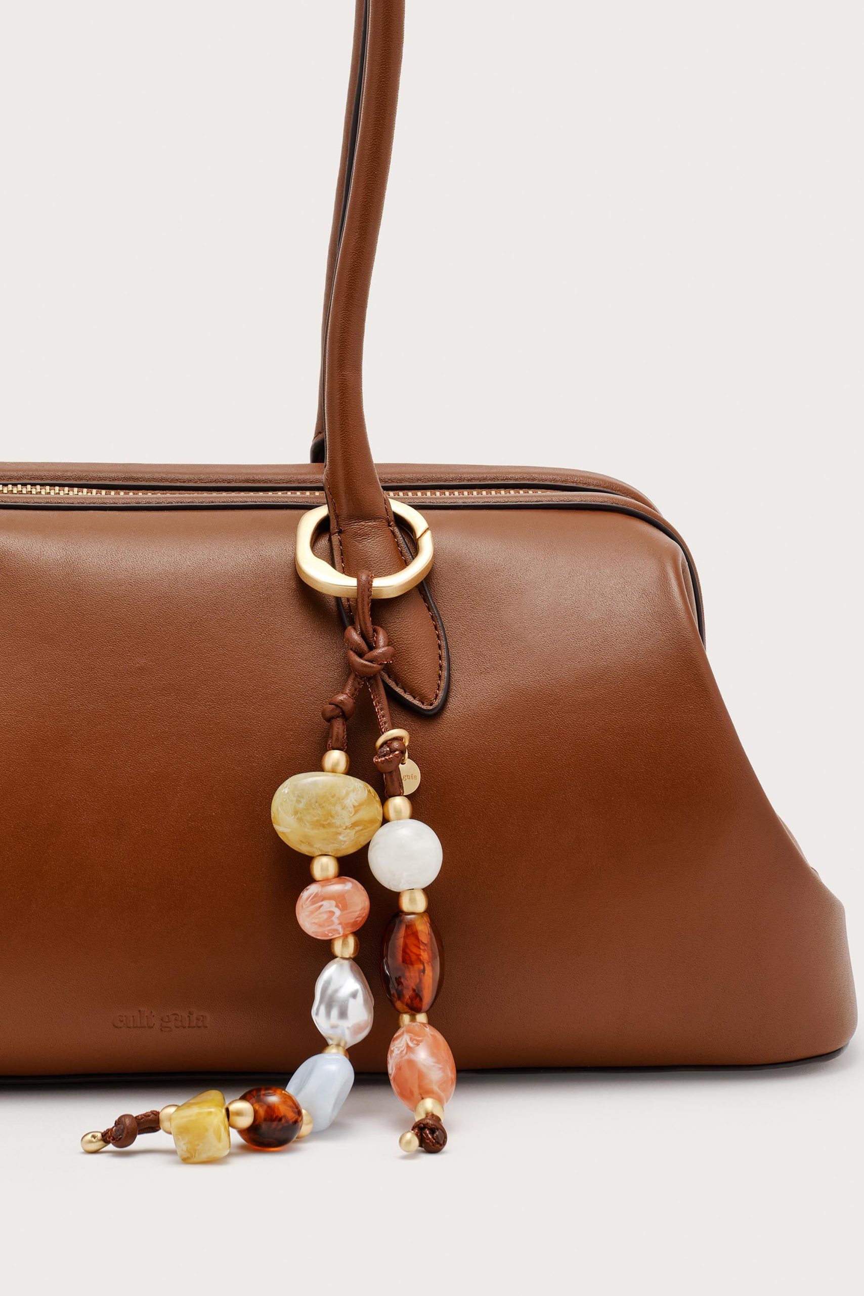 IMG_1306 - New Trend Close-up of a brown leather handbag with a gold ring hardware and a beaded accessory hanging from the strap surface edge.